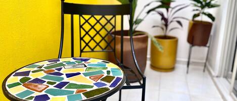 Una mesa pequeña y redonda con una cubierta de mosaico colorido, una silla de metal negra con respaldo de celosía y plantas en macetas frente a una pared amarilla.