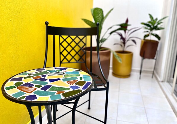 A small, round table with a colorful mosaic top, a black metal chair with a lattice backrest, and potted plants against a yellow wall. - Hotel Real Cocula (Cocula)