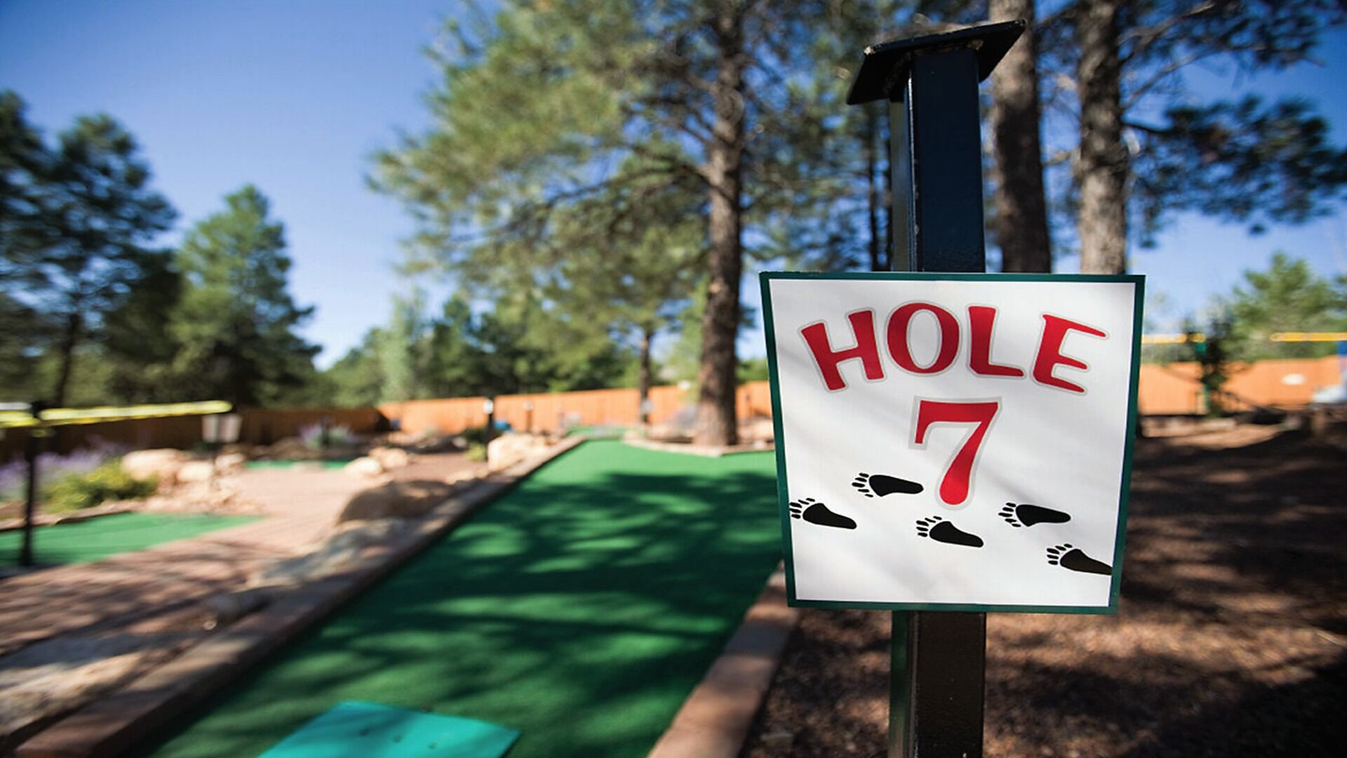 Een bord met 'HOLE 7', voetsporen en een achtergrond van een golfbaan met bomen en een heldere lucht.