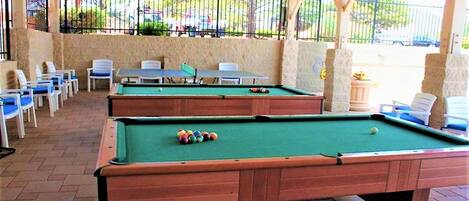 Two pool tables with chairs and a table in a covered outdoor area.