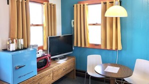 A room with a television, a small blue refrigerator, a wooden desk, two white chairs, and a table. - Hilltop Lodge Standard Queen - Room 105 (International Falls)