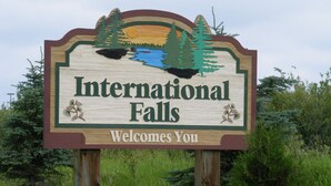 A wooden sign with "International Falls" and "Welcomes You" text, surrounded by greenery.