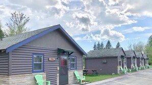 A row of log cabins with a sign reading "WILLOW" on the first cabin, green plastic chairs outside, and a parking lot in the foreground.