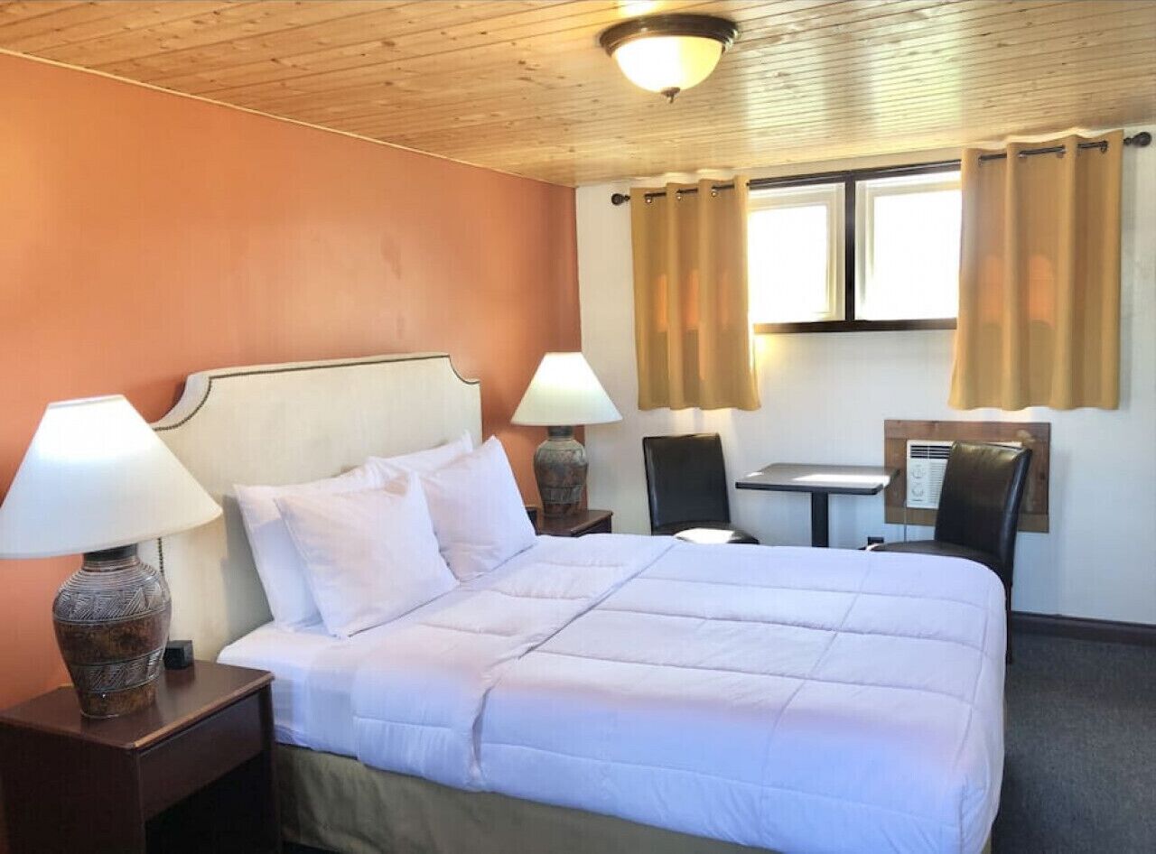 Une chambre d’hôtel avec un lit, deux lampes de chevet, une petite table, une chaise et une fenêtre avec des rideaux.