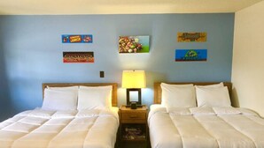 A hotel room with two beds, a nightstand with a lamp, and wall decorations including game-themed posters. - Hilltop Lodge Standard Double - Room 110 (International Falls)