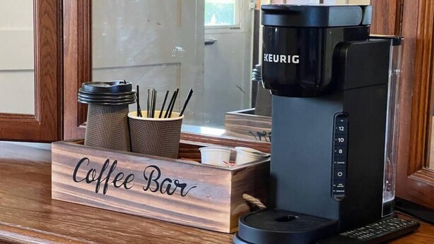 Una cafetera, un control remoto y un letrero de cafetería sobre una encimera de madera.