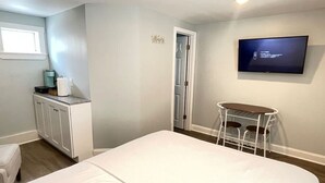 A bedroom with a bed, a small table with chairs, a TV on the wall, and a door leading to another room. - Hampton Beach Getaway | Mins to Portsmouth+ Maine (Hampton)