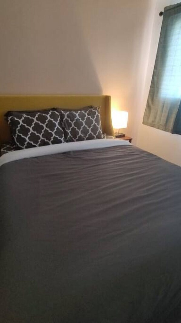 A bed with a dark grey sheet and a patterned pillow, a bedside lamp, and a window with a curtain.
