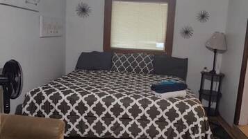 A bedroom with a bed, a chair, a window with blinds, a lamp, and a wall clock.