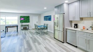 A modern kitchen with grey cabinets, a stainless steel refrigerator, and a dining area with a table and chairs. - Beachside Retreat | Hot Tub | Fire Pit | WiFi (West Palm Beach)