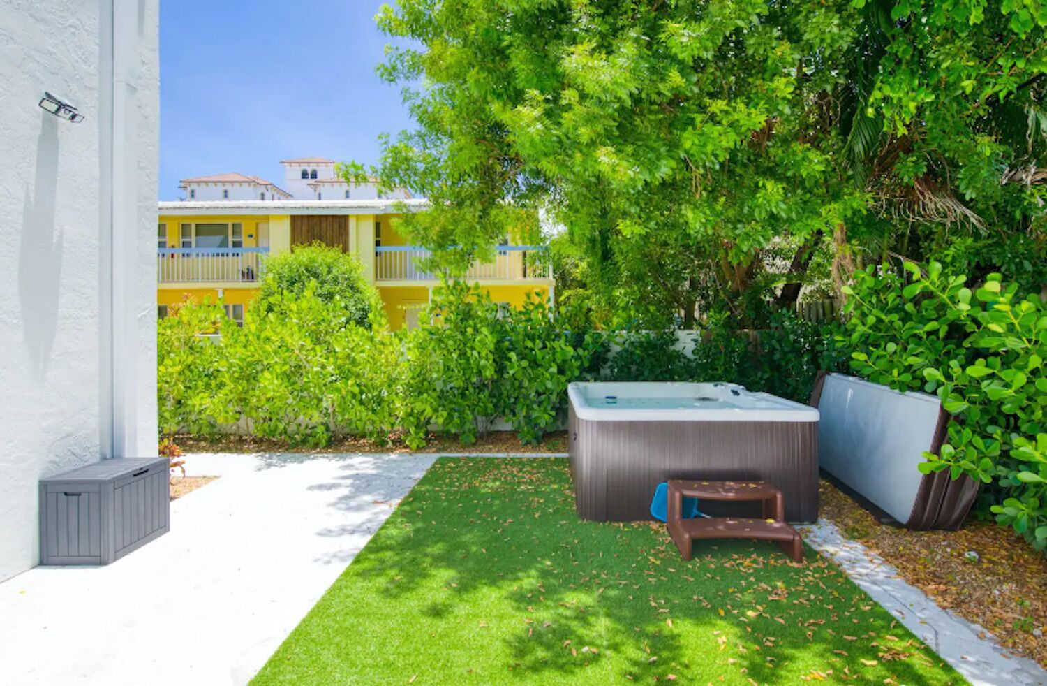 A backyard with a hot tub, a small table, and a bench. There is a white wall and a yellow building in the background.