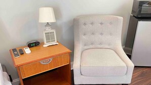 A small wooden nightstand with a lamp, a digital clock, and a remote control. Next to it is a light-colored upholstered chair with a tufted backrest. - Hampton Beach Getaway | Mins to Maine | Portsmouth (Hampton)