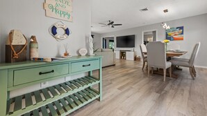 Een moderne woonkamer met een groene console tafel, een eethoek met grijze stoelen, een aan de muur bevestigde televisie en een plafondventilator.