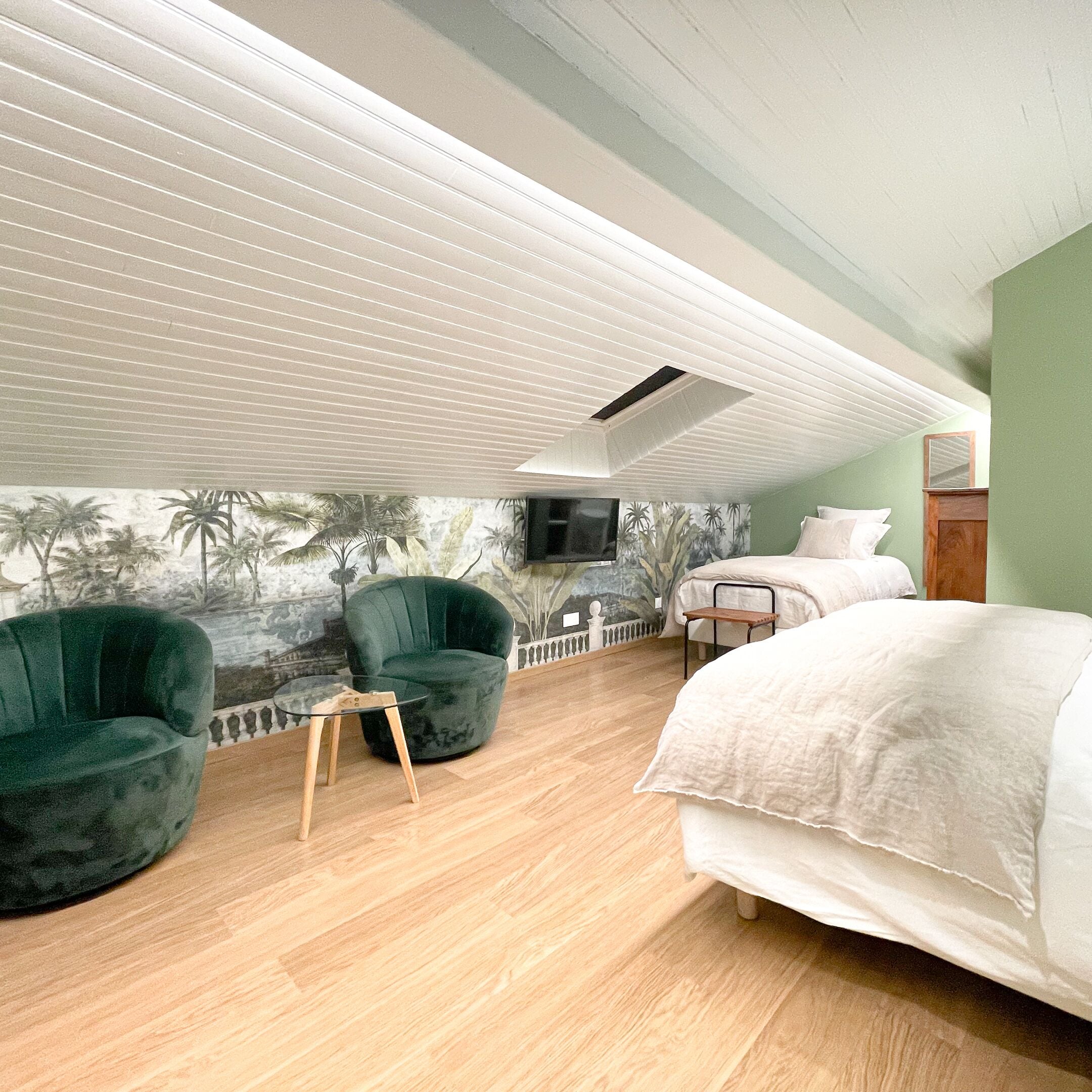 A bedroom with a bed, a chair, a small table, a TV mounted on the wall, and a mural of palm trees.
