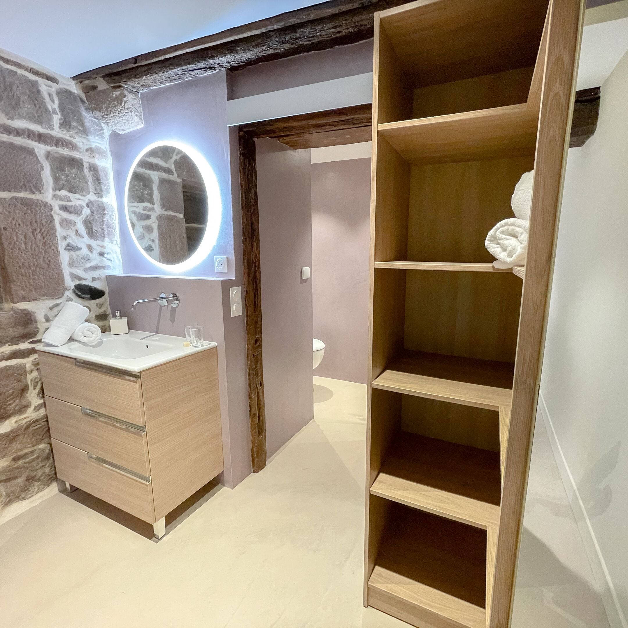 Un baño con tocador de madera, un espejo redondo con iluminación, un inodoro y una unidad de estanterías de madera.