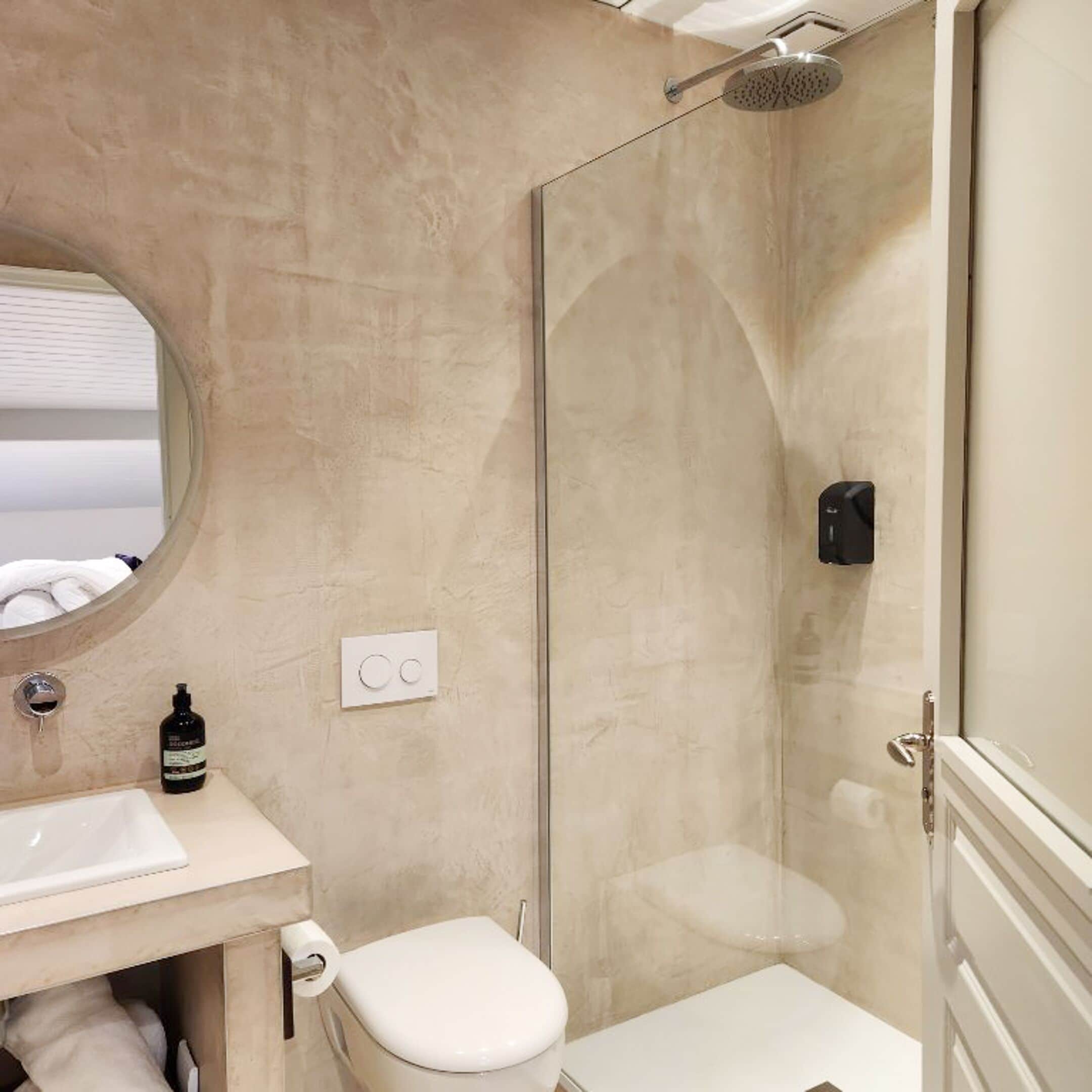 Een badkamer met een douche met glazen wand, een ronde spiegel, een wit wastafelmeubel en een toilet.