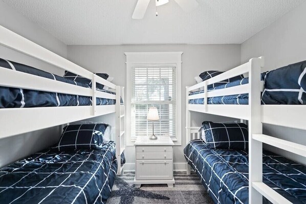 A room with bunk beds, a small white nightstand, and a window with blinds.