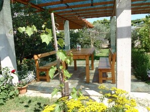 Zona d'estar a l'aire lliure coberta, amb taula de fusta i bancs, envoltada de plantes i flors en testos.