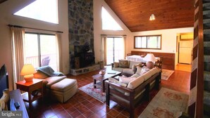 A spacious living room with a stone fireplace, wooden ceiling, and large windows.
