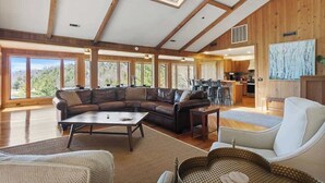 A spacious living area with a large sectional sofa, a coffee table, and a wicker side table. There is a kitchen in the background with wooden cabinets and a dining area.