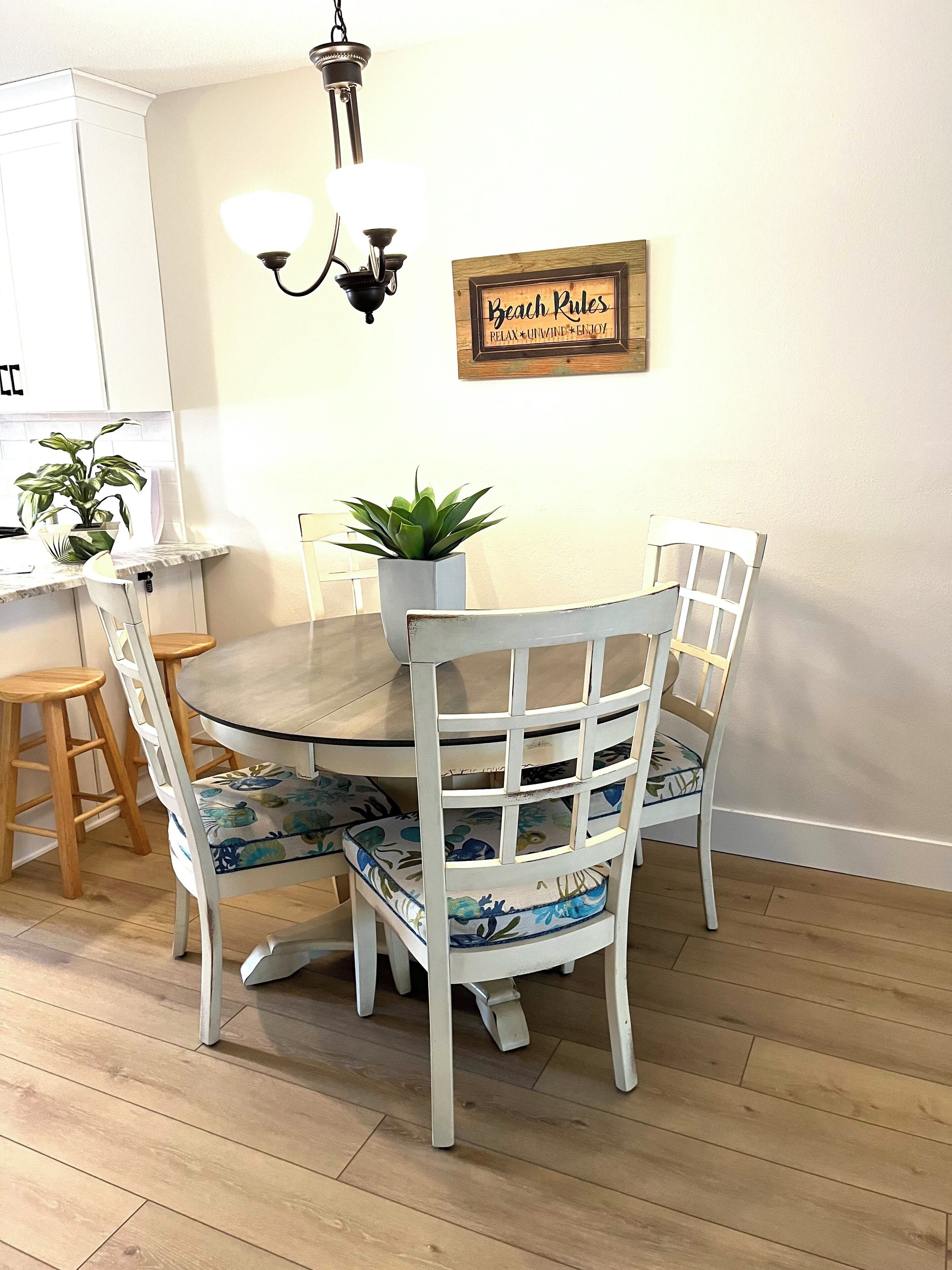 Un comedor con una mesa redonda, sillas blancas con tapicería floral, una planta en maceta y un letrero en la pared que dice "Beach Rules".