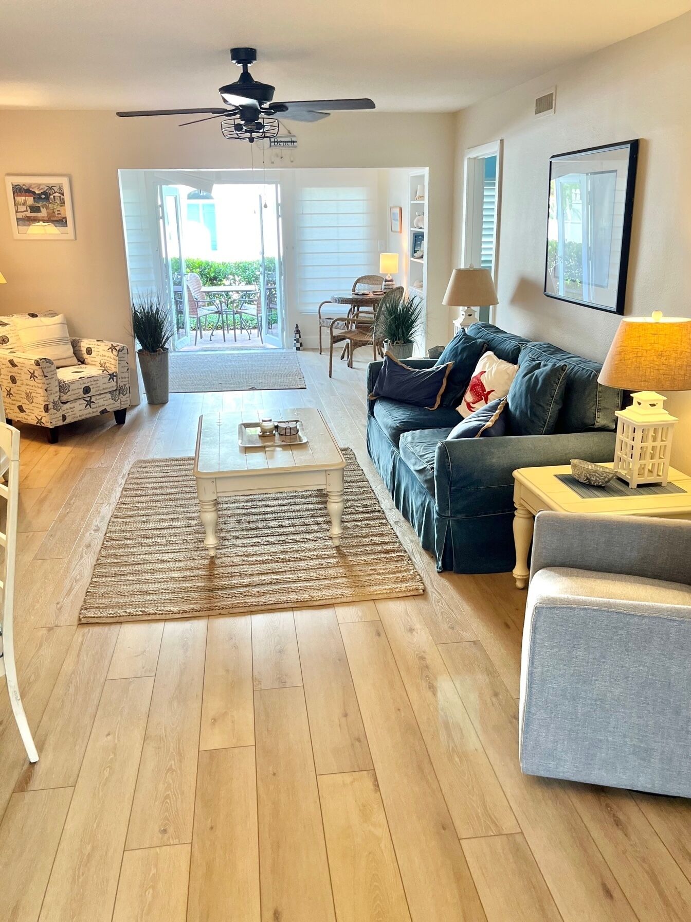 A spacious living room with a wooden floor, a ceiling fan, a sofa, a coffee table, and a view of the outdoors through a glass door.