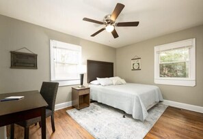 Ein Schlafzimmer mit einem Bett, einem Schreibtisch, einem Stuhl, einem Nachttisch, einem Deckenventilator und zwei Fenstern.