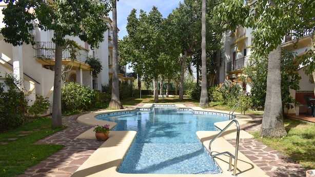 A swimming pool with a stone deck, surrounded by trees and a multi-story building with balconies. - Anacasa El Patio Denia PB1117 (Denia)