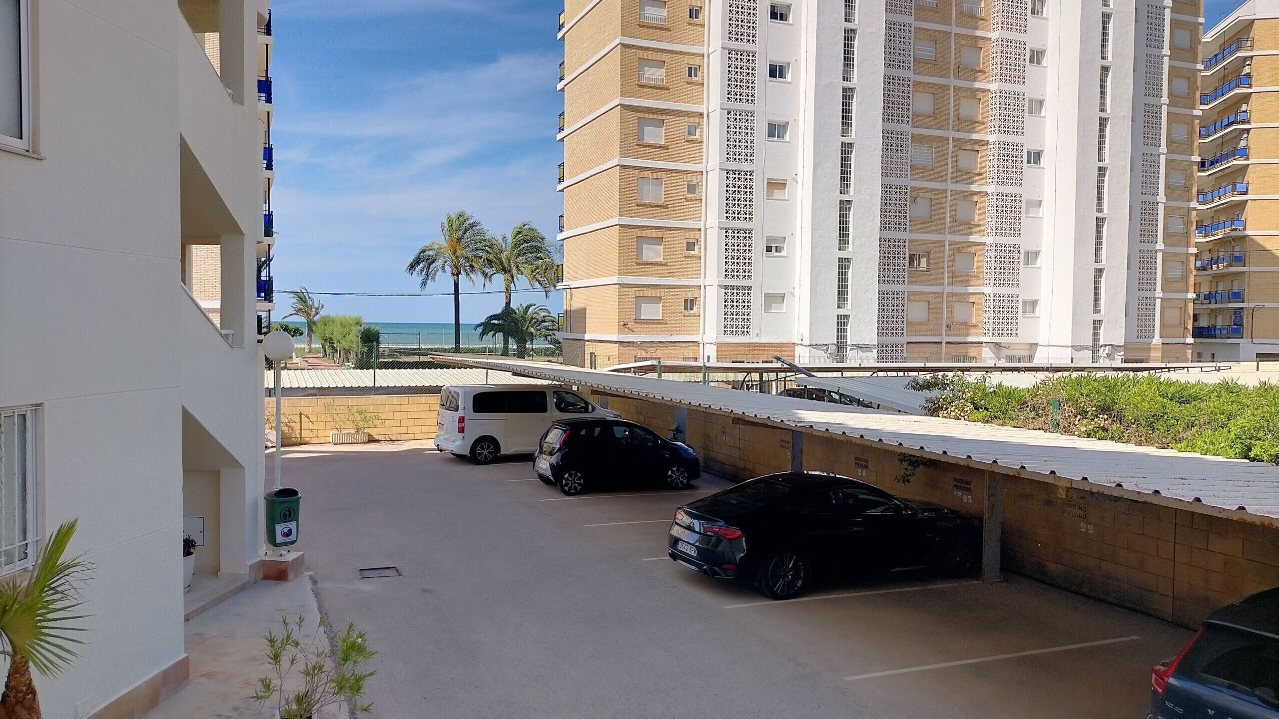 A parking area with several cars, a white van, and a building with multiple floors and balconies.