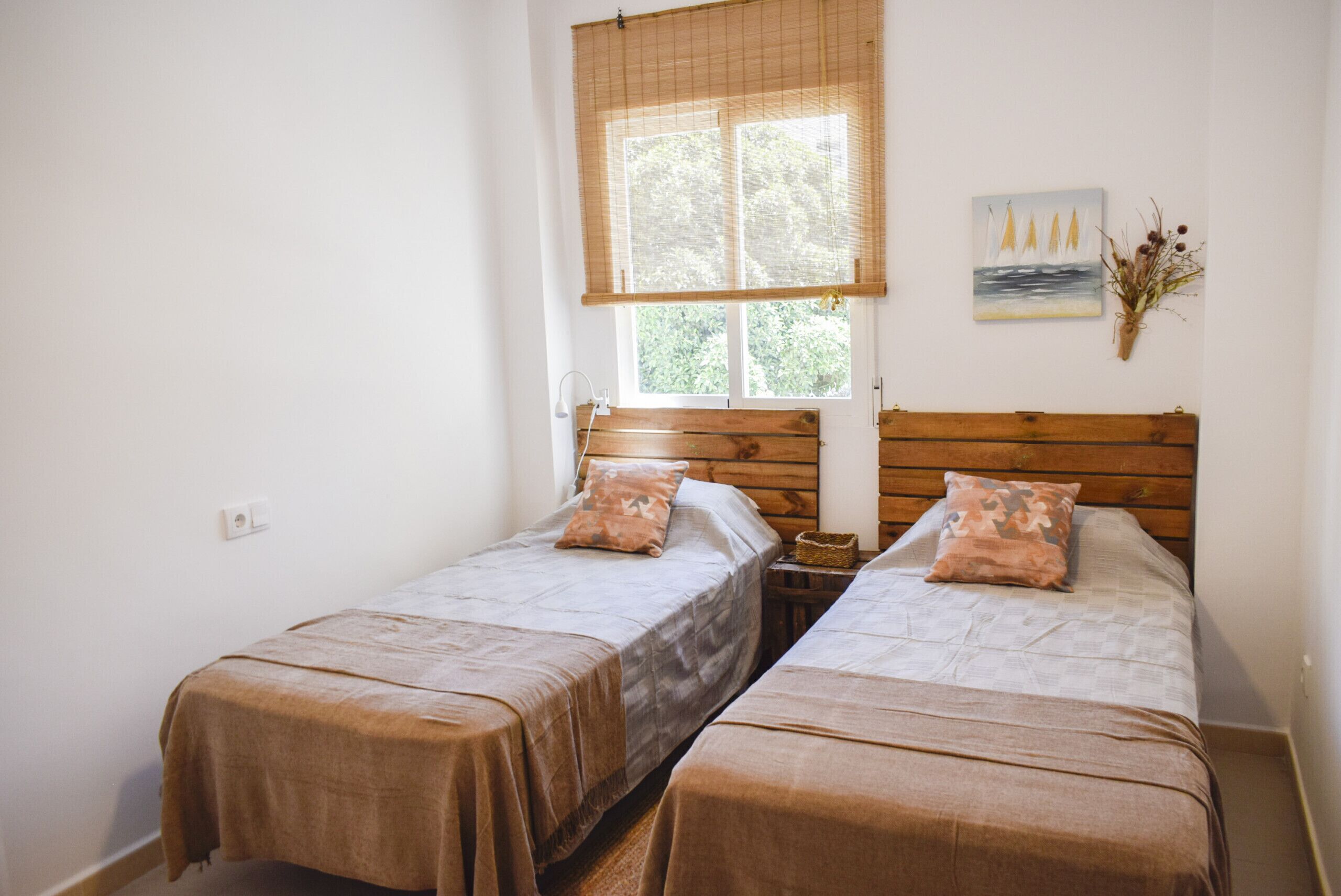 A room with two single beds, a window with a bamboo blind, a wall-mounted painting, and a vase with dried flowers.