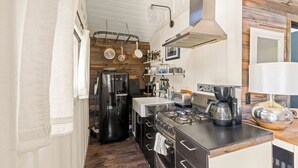 A compact kitchen with a stove, oven, and coffee maker. There is a wooden wall and a hanging pot rack. - The Arrow Cottage (Bend)