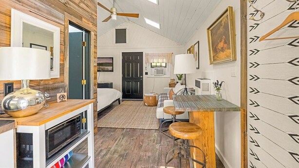 A compact hotel room with a kitchenette, a bed, a dining area with stools, and a ceiling fan. - The Arrow Cottage (Bend)