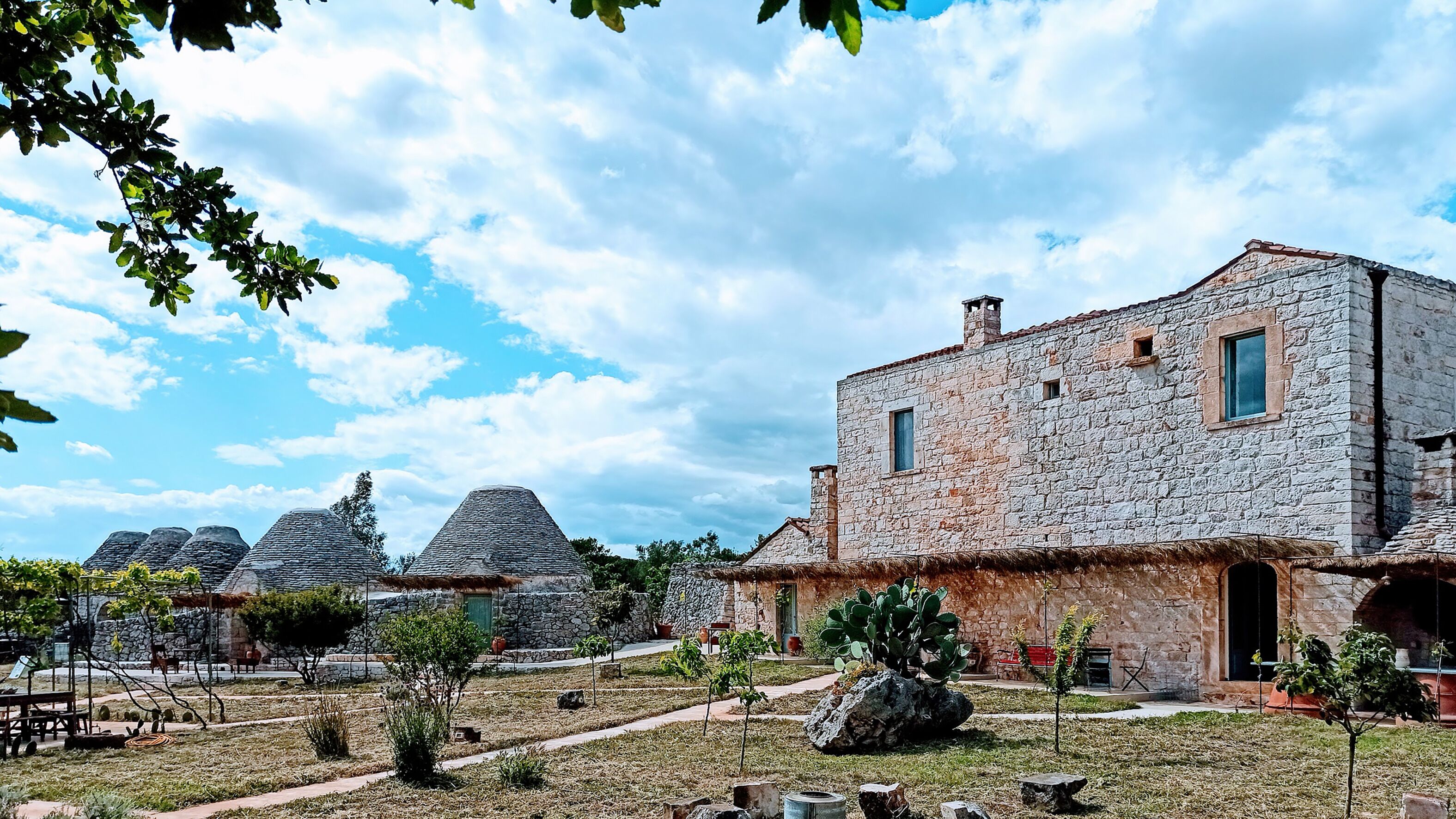 Un edificio de piedra con techo de paja, rodeado de un jardín con árboles pequeños y cielo despejado.