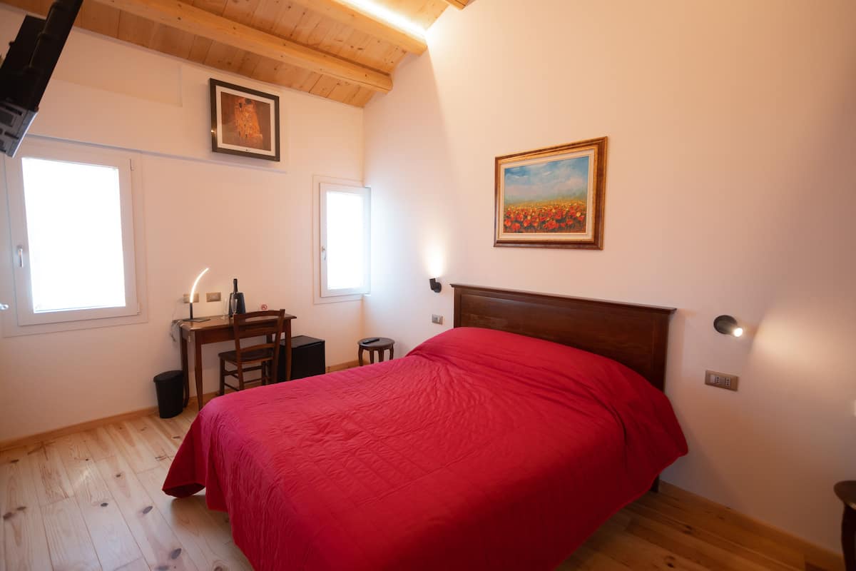Ein Schlafzimmer mit Holzbalkendecke, einem Bett mit roter Decke, einem Schreibtisch mit Stuhl und zwei Fenstern.