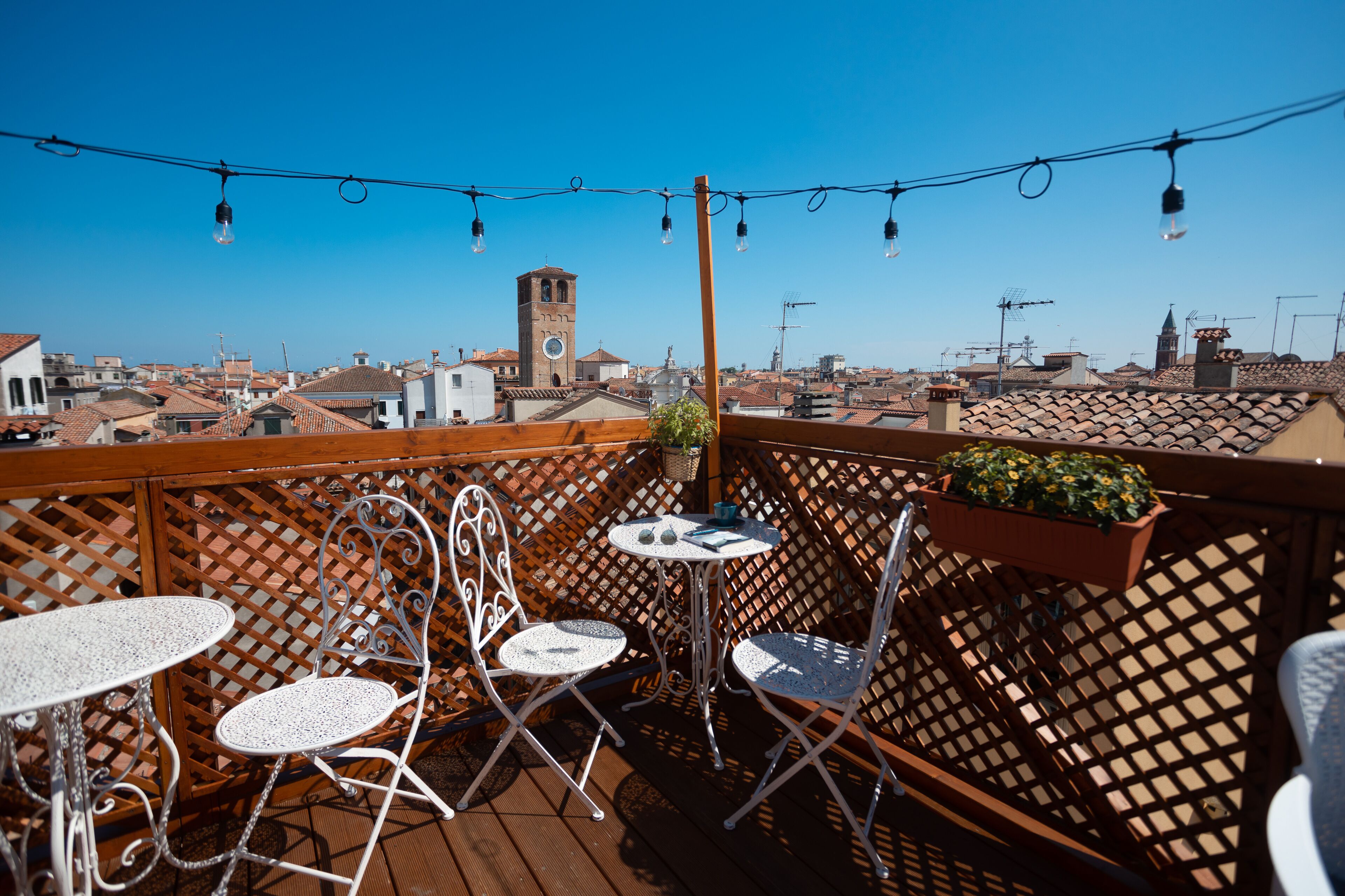 Una terraza en la azotea con cubierta de madera, sillas metálicas, una mesita y guirnaldas de luces.
