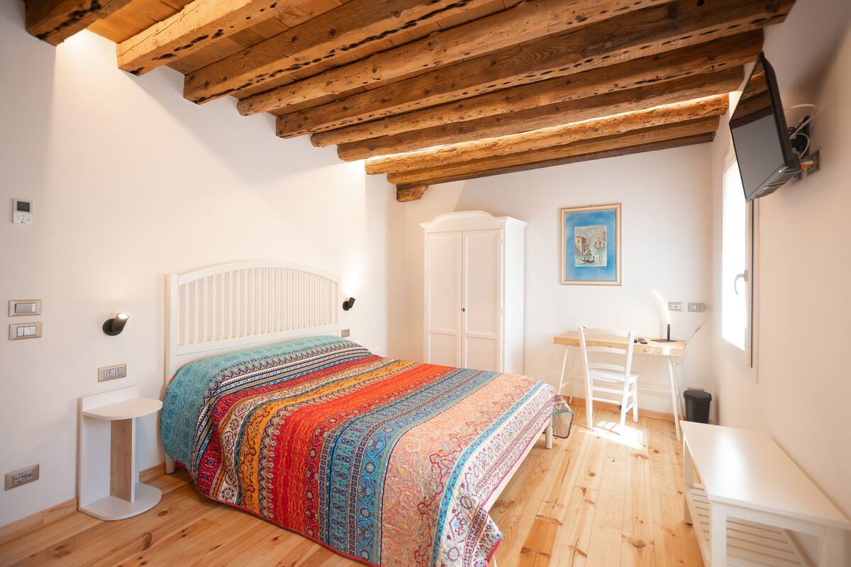 Ein Schlafzimmer mit Holzboden, einem Bett mit farbenfroher Decke, einem weißen Kleiderschrank, einem Schreibtisch mit Stuhl und einem an der Wand montierten Fernseher.