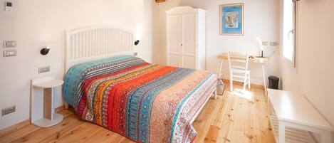 A bedroom with a wooden ceiling, a bed with a colorful blanket, a white wardrobe, a desk with a chair, and a TV mounted on the wall.