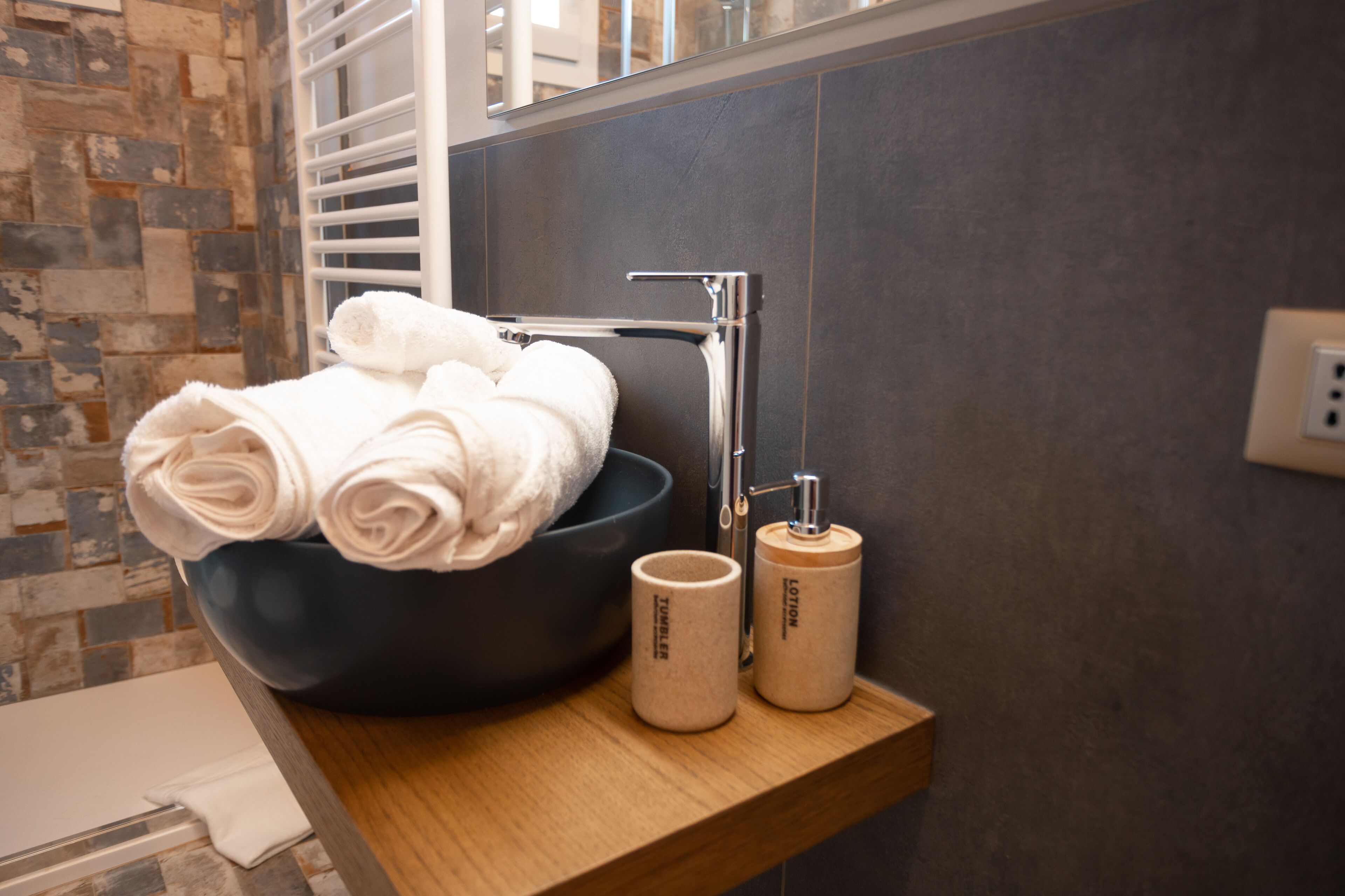 A modern bathroom with a large dark bowl sink, two beige toiletry containers, and neatly folded white towels on a wooden shelf.