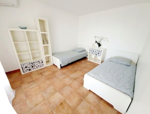 A small, clean room with two single beds, a white wardrobe, and a small cabinet with decorative tiles.
