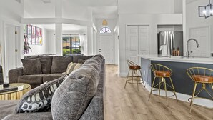 A spacious living area with a grey sofa, a round coffee table, and a modern kitchen with a stainless steel refrigerator and bar stools. - Luxury Corner Home, Arcade, Heated Pool & Spa. No extra fees. Pet Friendly. (Davenport)