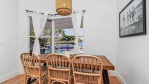Zona de menjador amb taula de fusta i cadires de vímet, lluminària penjant i vistes a una piscina i palmeres a través d'una finestra amb cortines blanques.