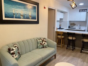 Un salon moderne avec un canapé bleu clair, un tableau représentant des voiliers et un coin cuisine doté de meubles blancs et de tabourets de bar.