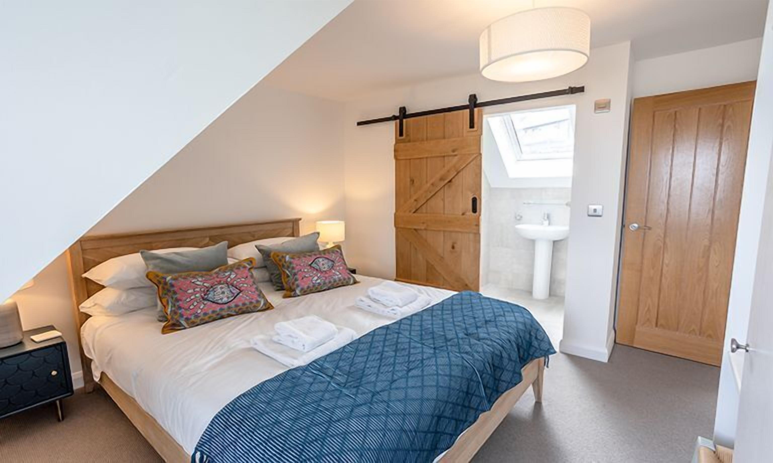 A bedroom with a sloped ceiling, a bed with a blue quilt, two pillows, a nightstand, a lamp, a wooden wardrobe, and a bathroom with a sink and a door.