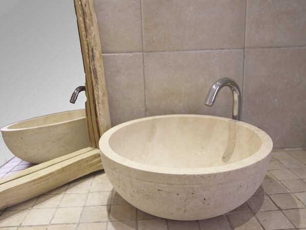 A round, beige ceramic sink with a single faucet, a wooden-framed mirror, and tiled walls.