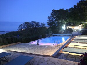 An outdoor swimming pool with a stone-tiled deck, lounge chairs, and a view of the sea.