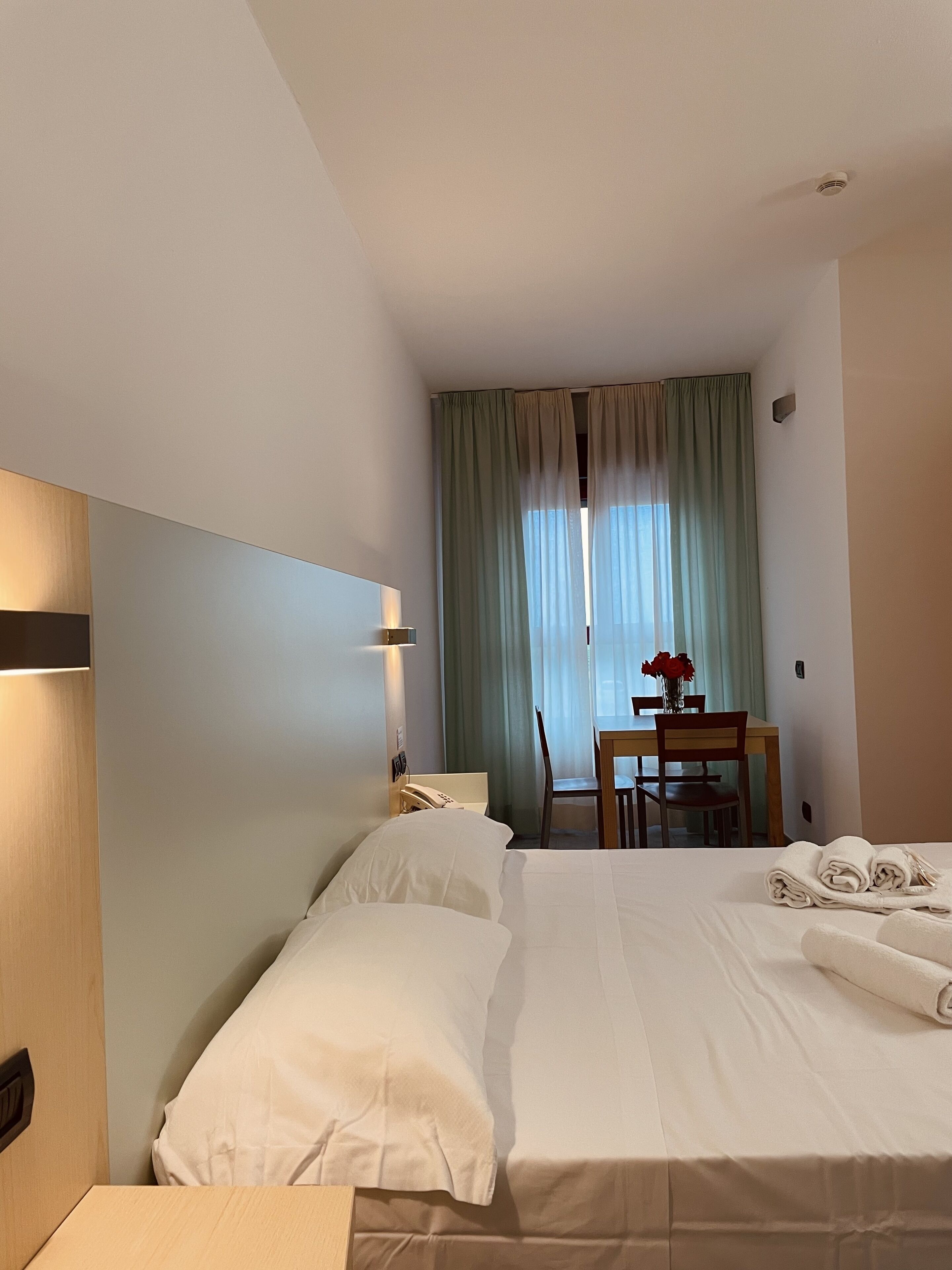 A hotel room with a bed, a desk, and a chair. There is a window with curtains, a bedside lamp, and a vase with flowers on the desk.