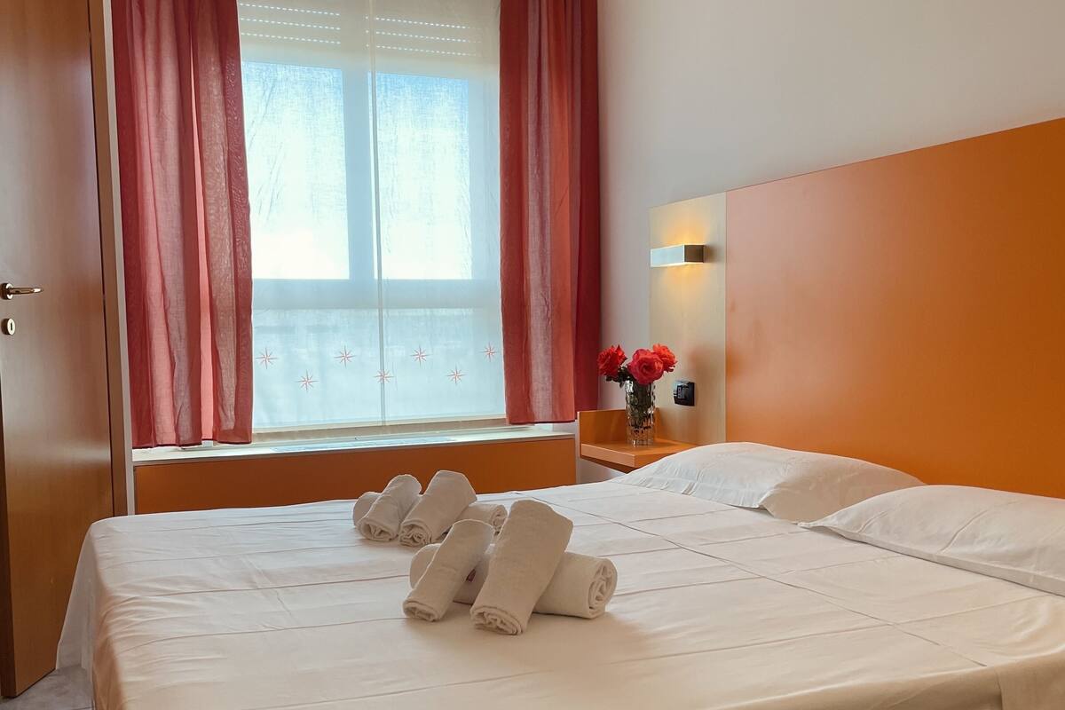 Une chambre d’hôtel avec un grand lit, une fenêtre avec des rideaux, une table de chevet ornée d’un vase de fleurs et une lampe fixée au mur.