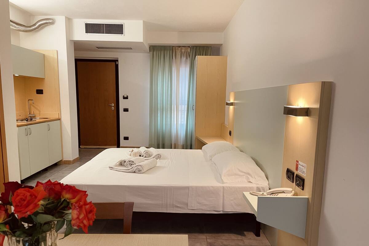 Une chambre d’hôtel comprenant un lit, une table de chevet, un téléphone, un miroir, un bureau avec une chaise, un vase avec des fleurs et une salle de bain avec un lavabo et des toilettes.