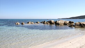 A beach with clear water, rocks, and a distant hill. - Apartment-Standard-Ensuite with Shower-Sea View-T3 (Porto-Vecchio)