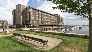 A waterfront building with a brick facade, a grassy area with a bench, and a marina with boats.